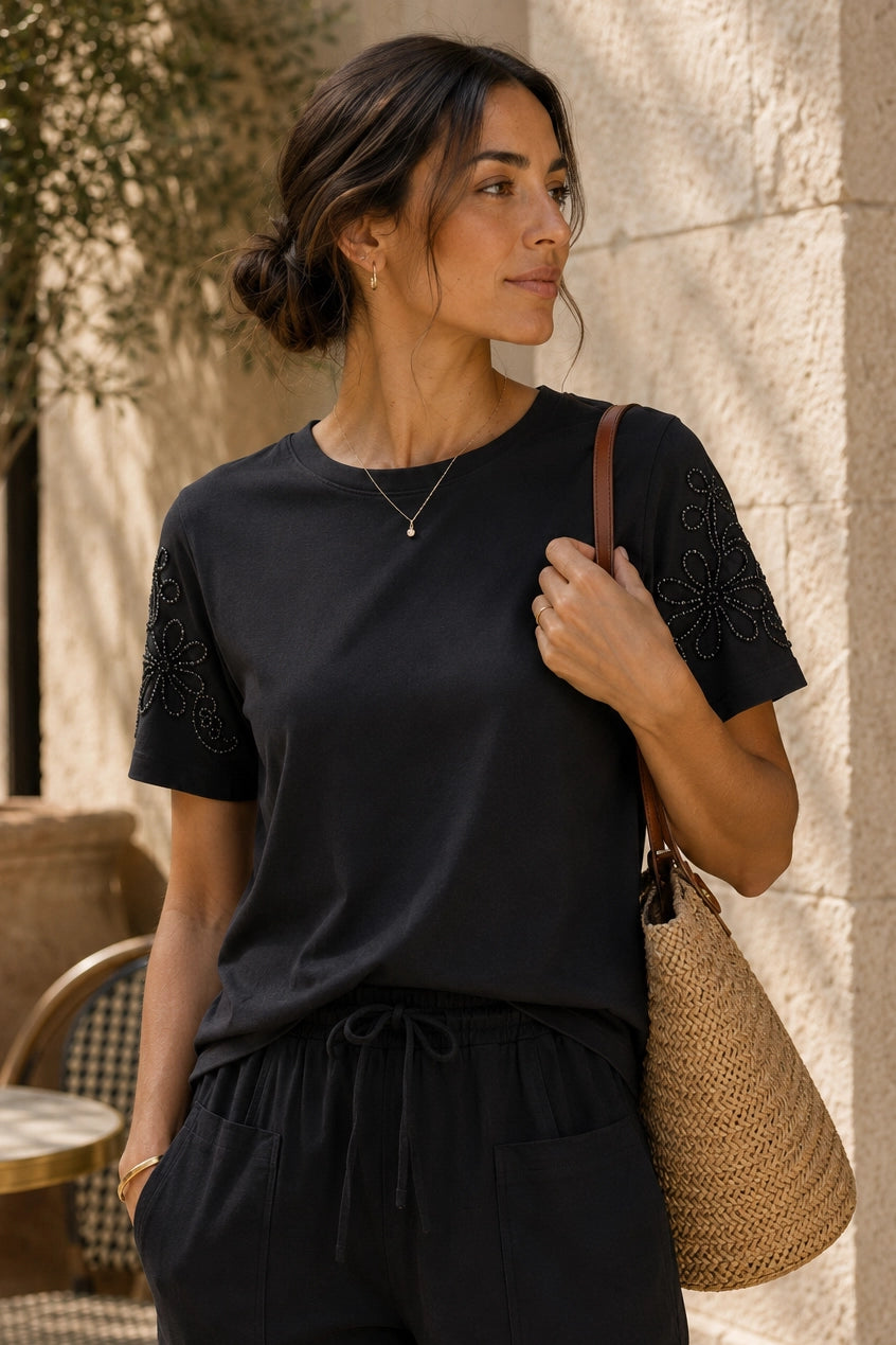 A woman wearing a black short-sleeve top with floral embroidery on the sleeves, paired with black drawstring pants and carrying a woven straw shoulder bag, standing against a light stone wall.