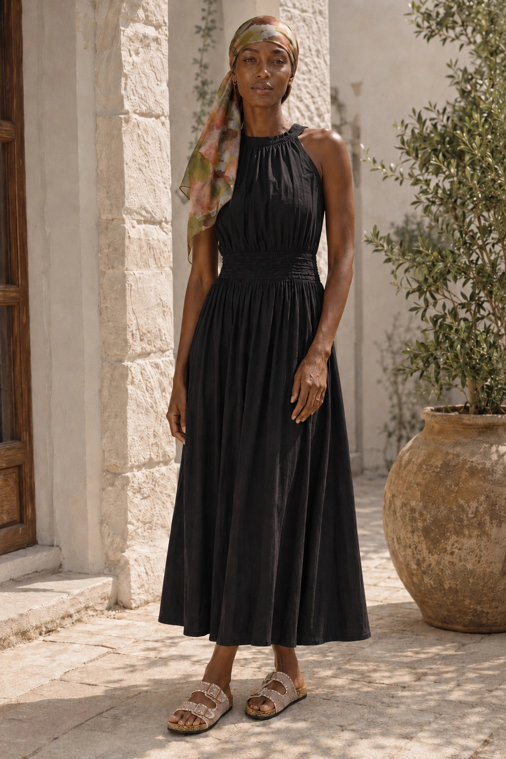 A woman wearing a sleeveless black midi dress with a gathered waist and a patterned headscarf, standing outdoors in front of a light stone wall and potted plants.