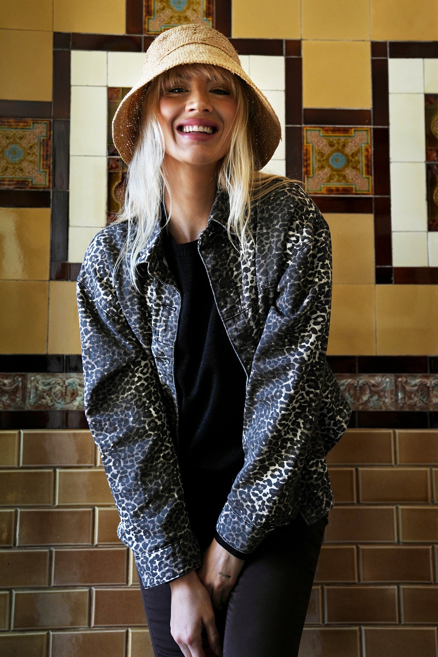 Woman wearing a patterned jacket and hat in front of a tiled wall.