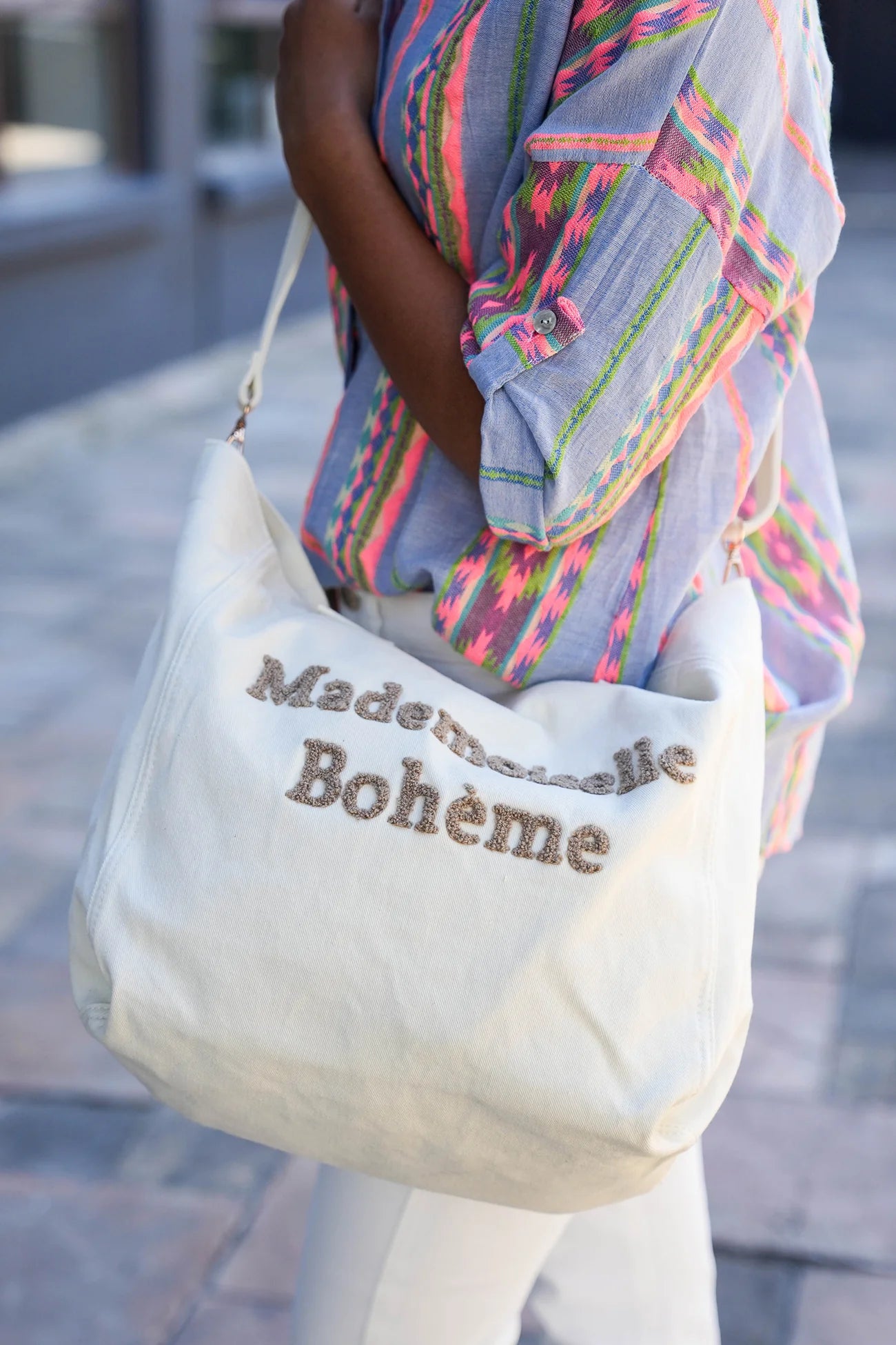 Person holding a white tote bag with 'Made in Bohème' text, wearing a colorful shirt.