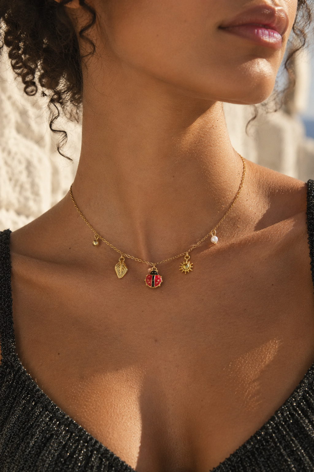A close-up lifestyle shot of a model wearing a delicate gold charm necklace. The necklace features a small red and black ladybug charm, a gold leaf charm, a gold star charm, and a tiny pearl bead, shown against the model's neck and upper chest.