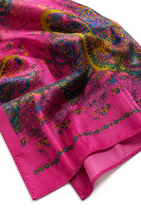 A close-up of a folded silk scarf featuring a vibrant magenta pink base with an intricate multicolor paisley pattern in yellow, blue, green, and purple, finished with a matching solid pink border.