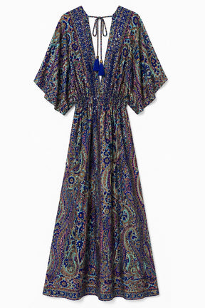A long kaftan-style maxi dress with a paisley print in purple, blue, green, and beige tones, featuring wide short sleeves, a deep V-neckline with a tie at the back, and a gathered waist.