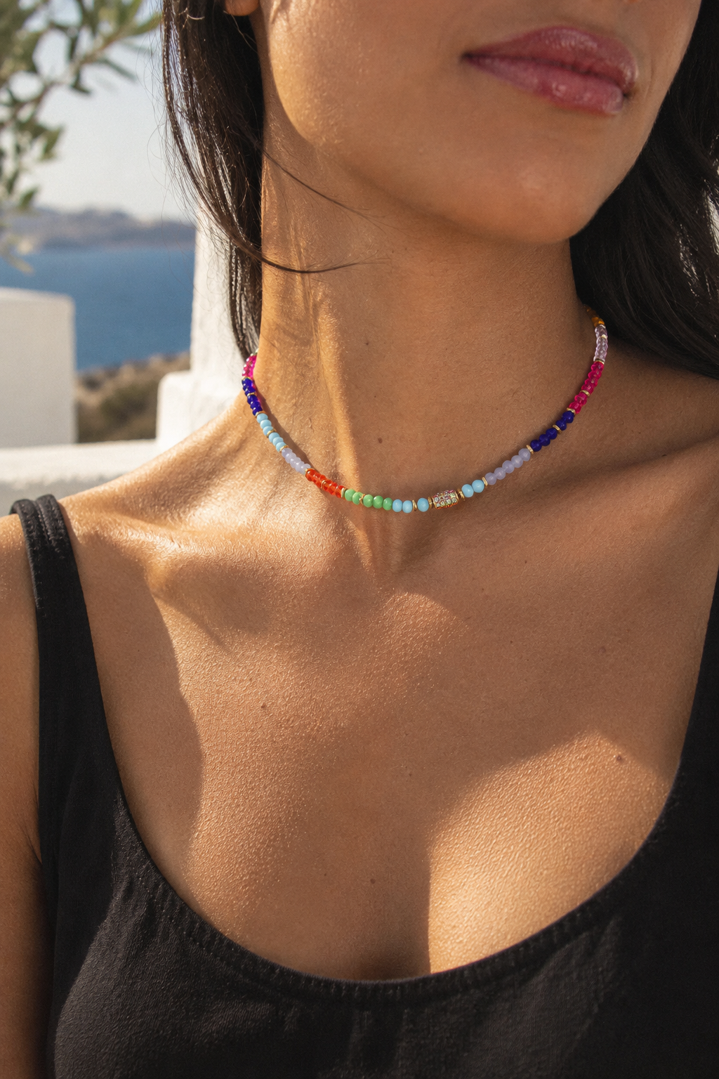 A close-up lifestyle shot of a woman wearing a colorful beaded choker necklace. The necklace features a gradient of beads from blue and red at the ends to green, yellow, orange, and light blue in the center, with a small white bead and gold heart charm in the middle.