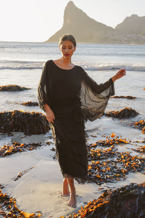 Woman in a black dress standing on a beach with mountains in the background