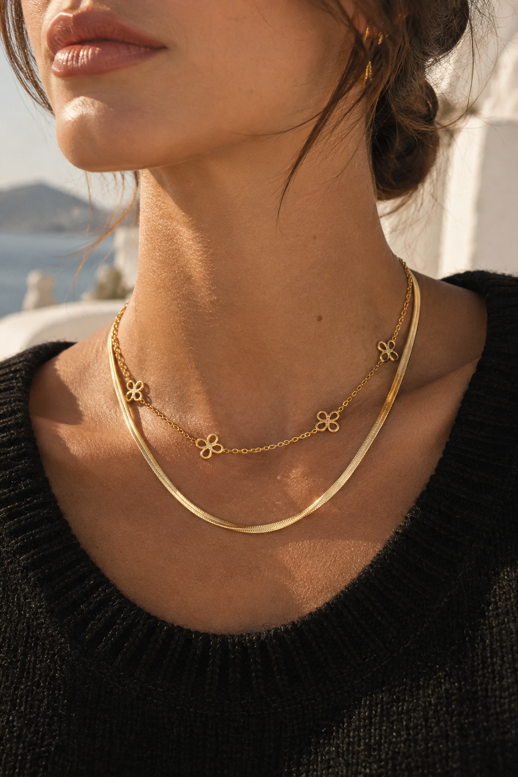 A close-up lifestyle shot of a model wearing a double-layered gold necklace. The necklace features a thin chain with small clover-like charms and a thicker, polished snake-style chain layered on top.
