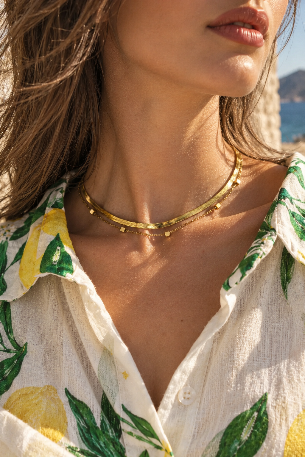A close-up lifestyle shot of a model wearing a delicate gold chain necklace adorned with small gold beads, styled with a white and green lemon-patterned blouse.