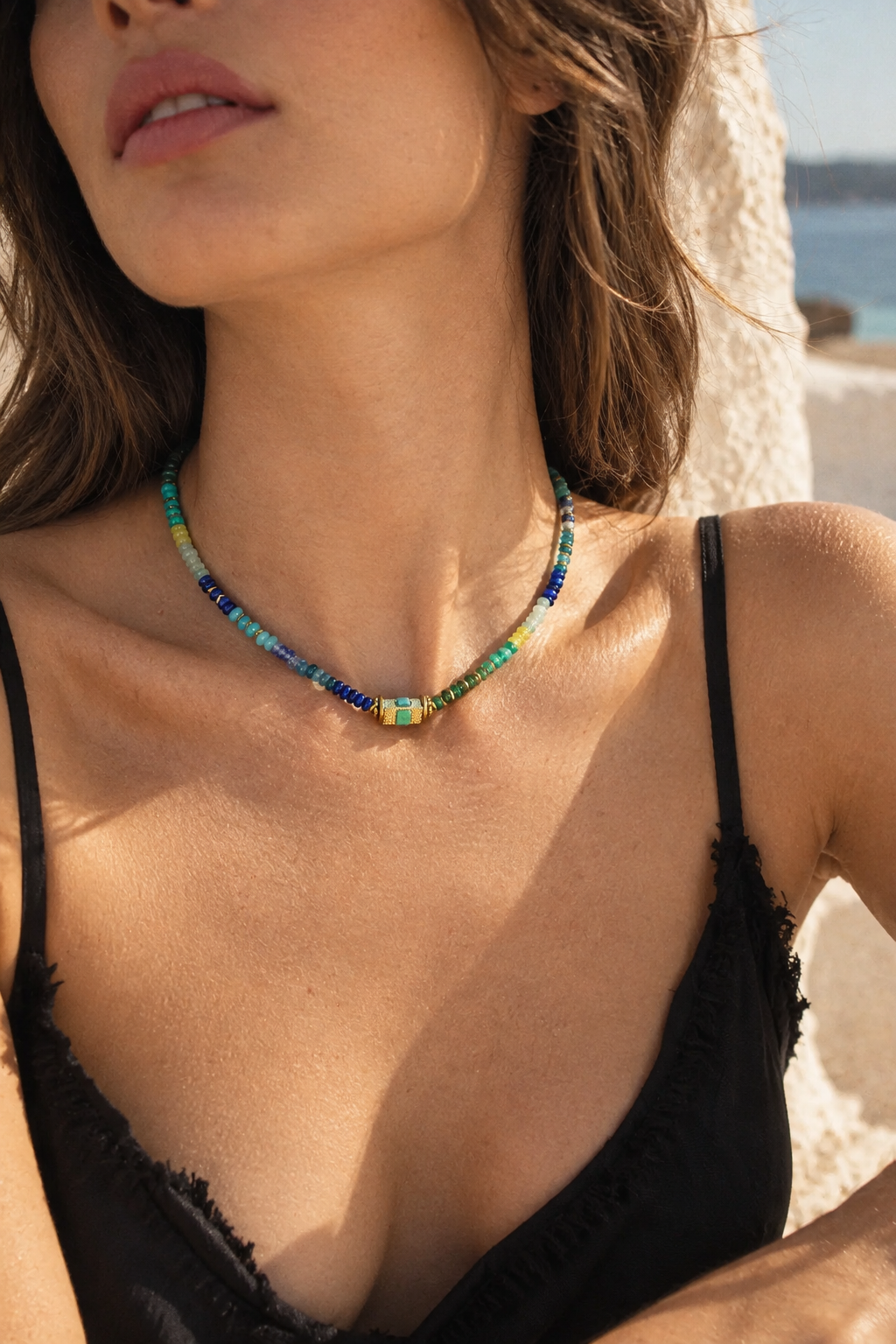 A close-up lifestyle shot of a woman wearing a colorful beaded necklace with a central yellow and green bead, set against a sunny beach background.