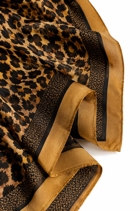 A close-up of a leopard-print scarf with a wide tan border and black patterned trim, laid out on a white background.
