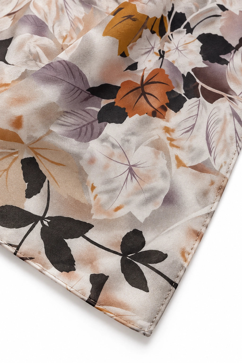 A close-up of a satin or silk scarf with a botanical floral print, featuring large leaves and flowers in black, purple, orange, and beige tones on a white base.
