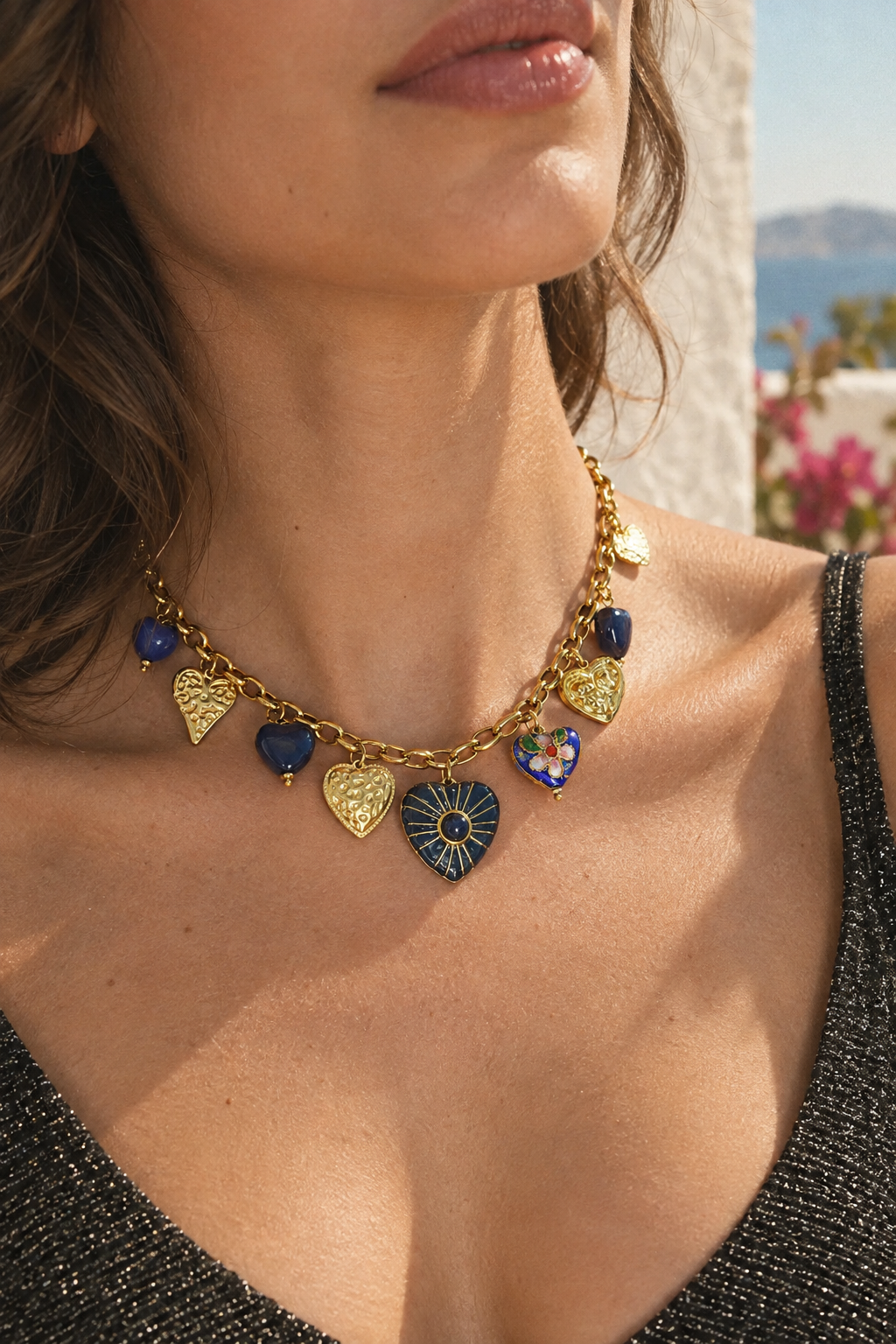A close-up lifestyle shot of a woman wearing a gold charm necklace with heart and sunburst charms in gold, blue, and multicolor enamel.