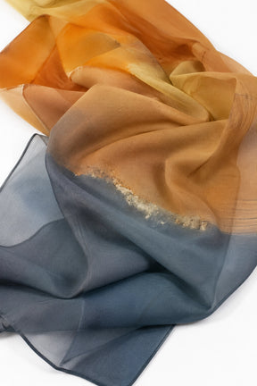 A lightweight sheer scarf draped in soft folds, featuring a gradient color story that transitions from warm golden orange at the top to a muted slate blue-gray at the bottom, with a subtle metallic shimmer along the center fold.