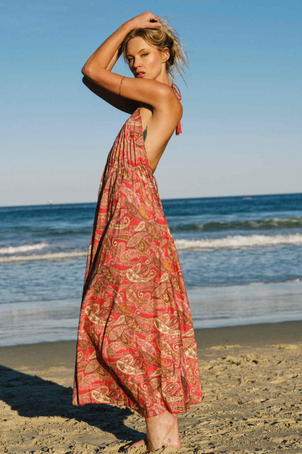 A woman stands on a sandy beach wearing a long, sleeveless halter dress with a red, orange, and gold paisley pattern. The ocean and blue sky are visible in the background.