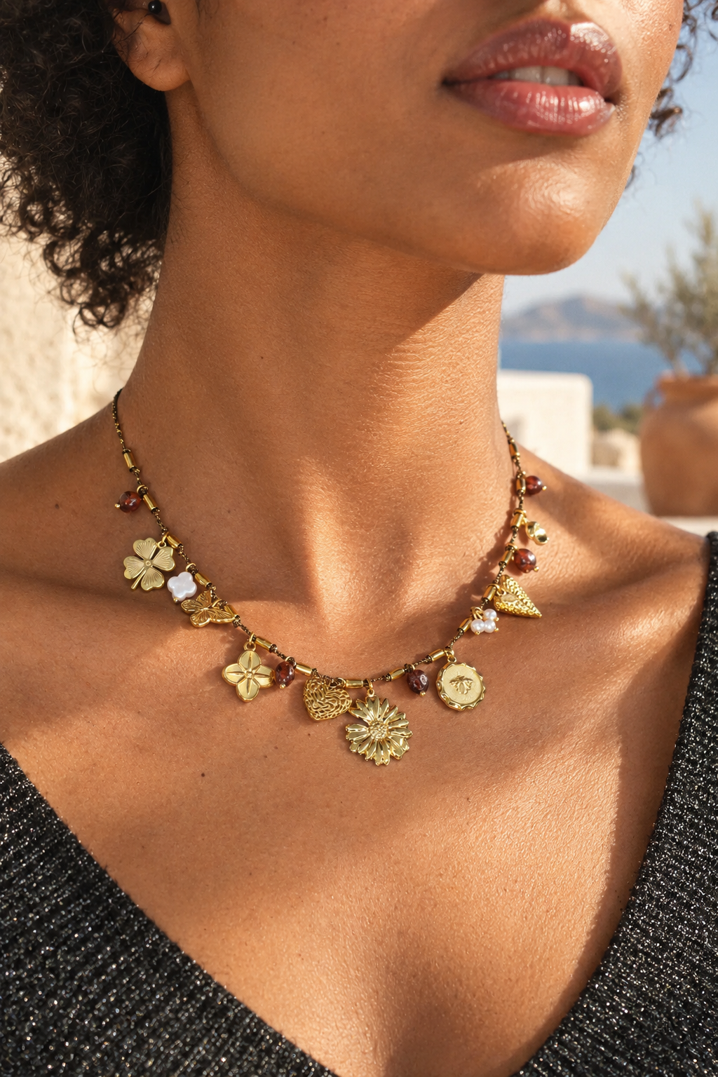 A close-up lifestyle shot of a model wearing a gold charm necklace with various organic-shaped charms, including a flower, leaf, and triangle, accented with small amber and white beads.