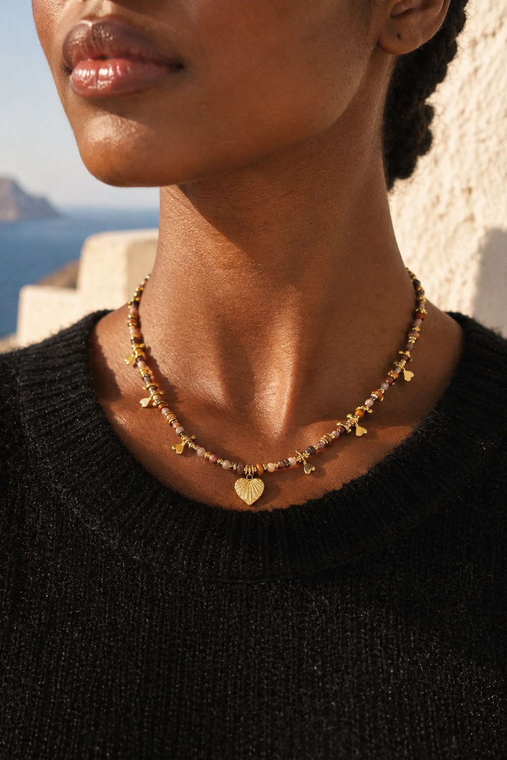 A close-up of a model wearing a delicate gold heart necklace with small gold charms and amber-colored beads, paired with a black knit sweater against a sunny outdoor background.