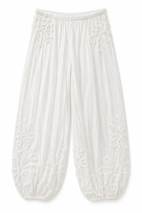 White crinkled cotton harem-style pants with embroidered floral details on the upper thighs and lower legs, featuring an elastic waistband and gathered cuffs.
