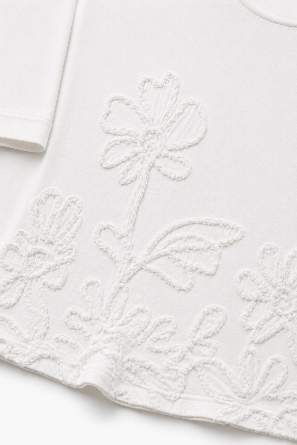 Close-up of a white short-sleeve top with tonal floral embroidery and textured applique along the hem.