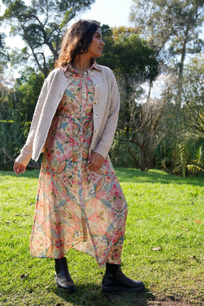 Woman in a floral dress and cardigan standing in a grassy field with trees in the background