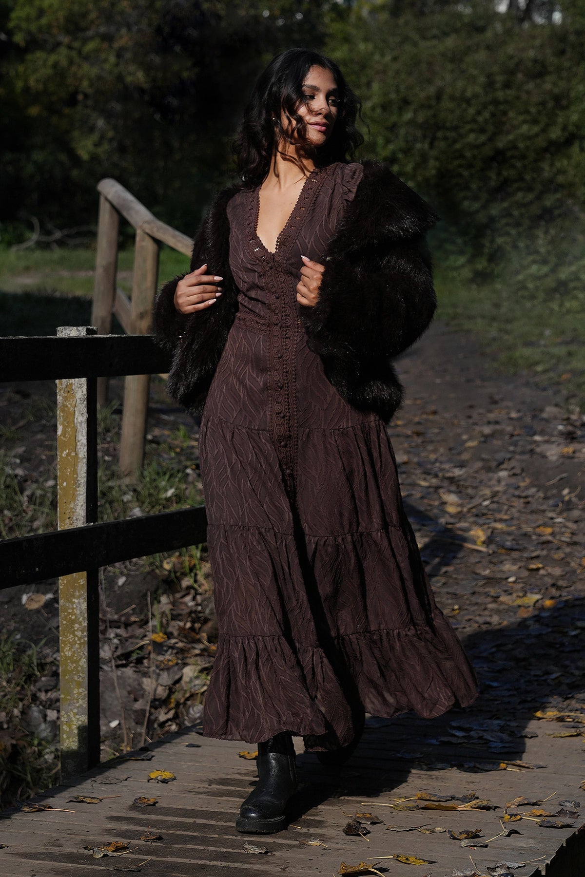 Woman in a long brown dress with a fur coat standing on a wooden path.