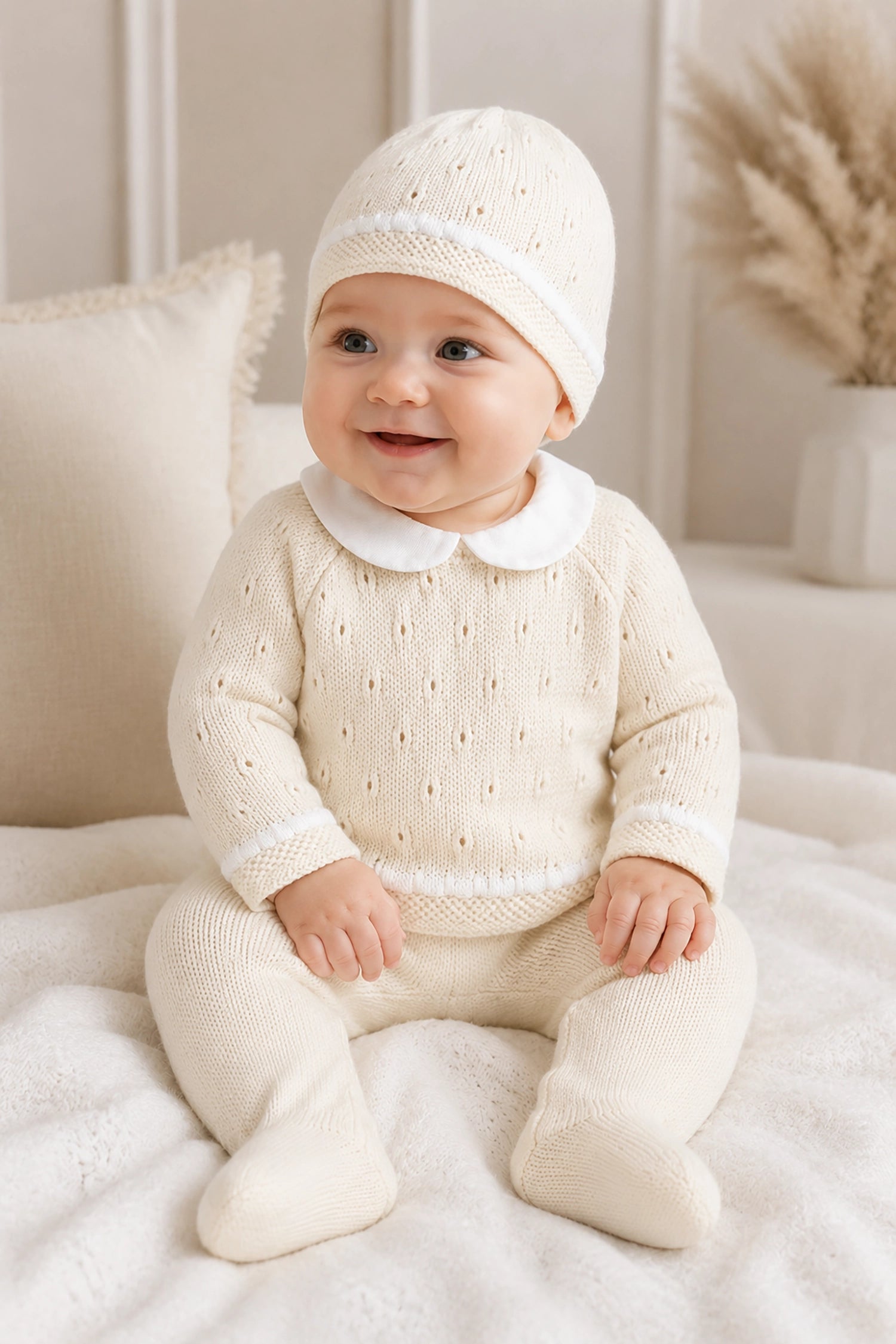 Baby in a white knit outfit sitting on a soft surface with a neutral background