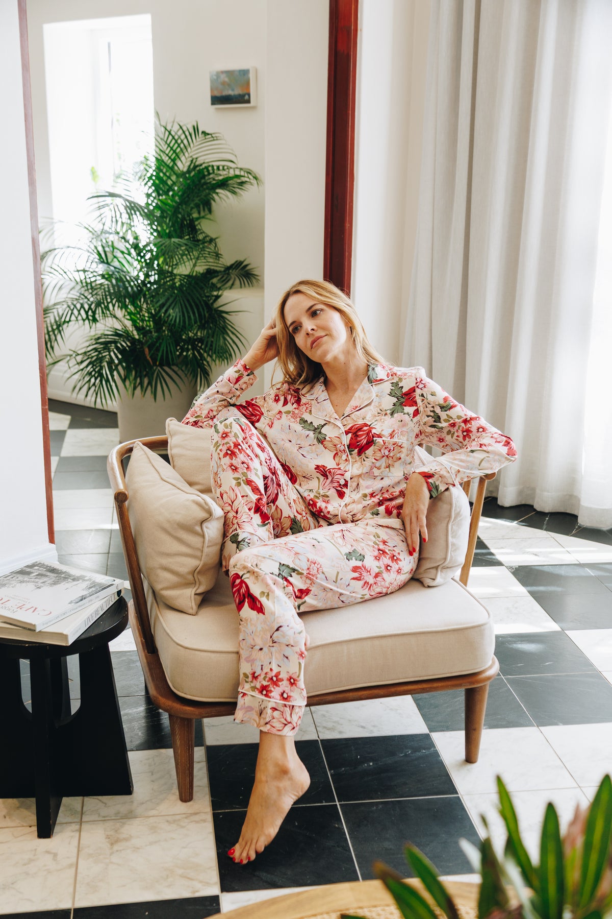 Woman in a floral robe sitting on a chair in a stylish room with plants and decor.