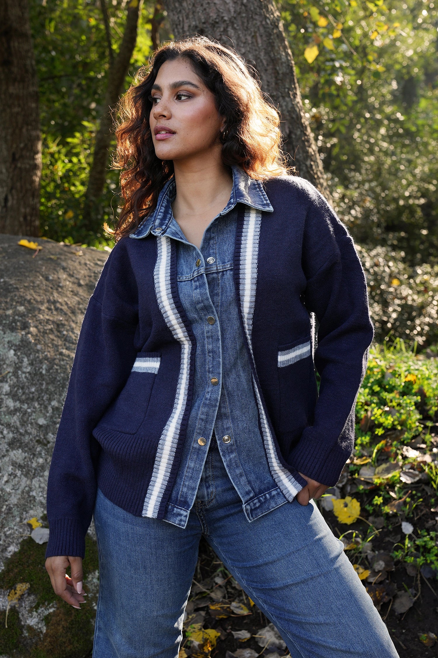 Woman wearing a blue cardigan with white trim standing outdoors near trees and rocks.