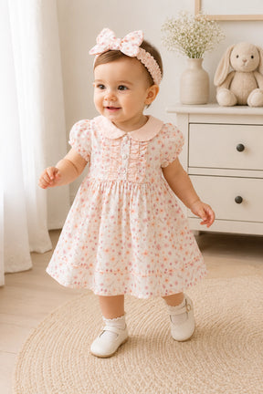 Baby girl in a floral dress standing in a room with a white dresser and decorative items.