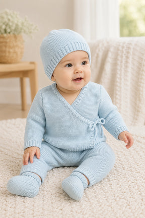 Baby wearing a light blue knitted outfit with matching hat on a soft surface.